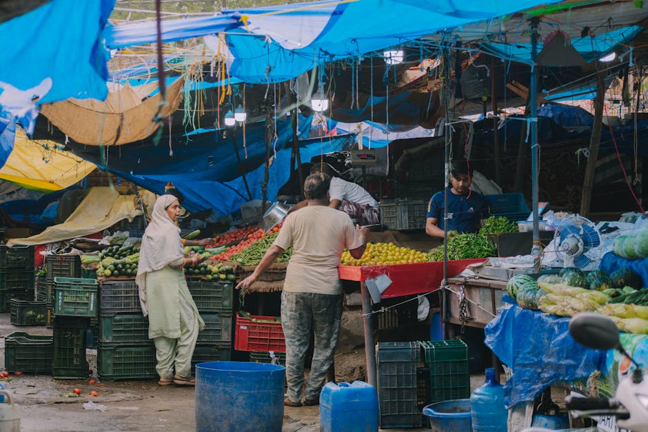 The image depicts a busy outdoor marketplace stall with a makeshift blue and yellow tarpaulin roof supported by wooden and metal poles, offering a variety of fresh fruits and vegetables. On the left,