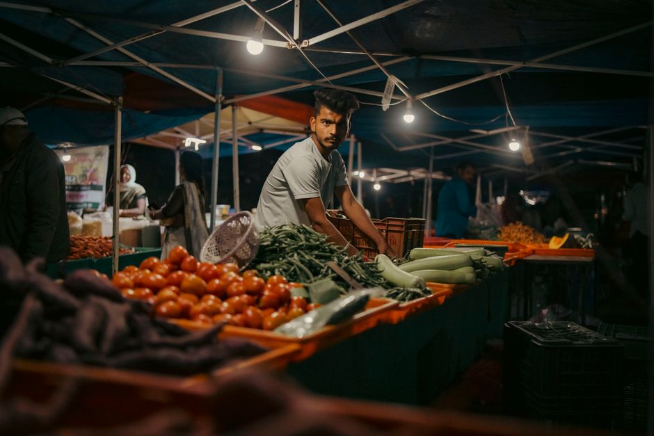 The image depicts a busy outdoor marketplace stall with a makeshift blue and yellow tarpaulin roof supported by wooden and metal poles, offering a variety of fresh fruits and vegetables. On the left,