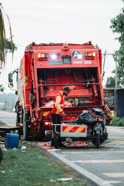 In this street scene, a large collection of mixed waste and rubbish bags is piled around multiple waste bins near a parking area. The waste includes black plastic bags, flattened cardboard boxes, plas